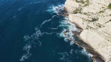 Dalgaların yukarıdan görünüşü mavi bir okyanusta kayalara çarpıyor. Deniz dalgaları güzel sahil manzaralı insansız hava aracı. Kuşların bakış açısı okyanus dalgalarının yukarıdan kayalıklara çarpmasını gösteriyor..