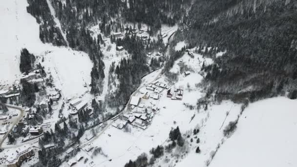 移动4K无人驾驶飞机飞越滑雪场村。空中冬季风景，在冰雪覆盖的森林和寒冷的山林之间有小村舍. — 图库视频影像