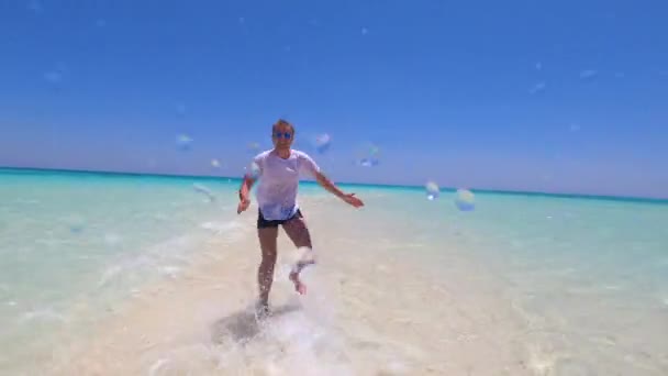 Un uomo con una maglietta bianca e occhiali corre lungo la riva dell'acqua turchese. Un ragazzo felice corre e schizza nell'acqua turchese dell'oceano. — Video Stock