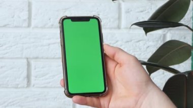 Man using smart phone in office space, sitting on chair and using green screen phone mockup, chroma key, browsing content, video, blogging, tapping center of screen, flipping through news flyer.