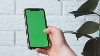 Man using smart phone in office space, sitting on chair and using green screen phone mockup, chroma key, browsing content, video, blogging, tapping center of screen, flipping through news flyer.