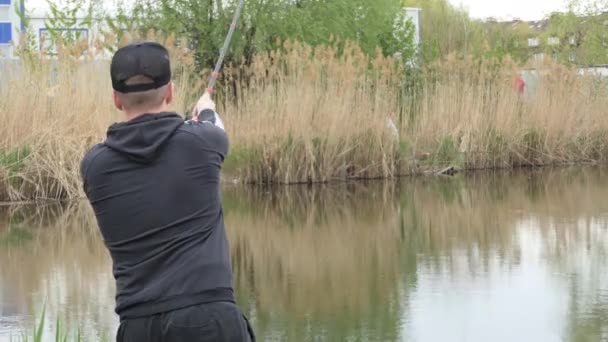 A man fishes, stands on the shore of a large and calm lake and catches fish with a fishing rod and spinning. The man threw the spinning rod into the calm water surface. — Stock Video