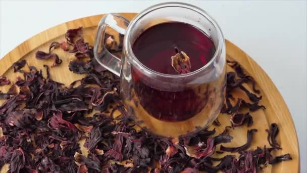 Κόκκινο τσάι σε γυάλινο κύπελλο σε λευκό φόντο. Κόκκινο τσάι ιβίσκος χύνεται σε ένα διαφανές ποτήρι με διπλά τοιχώματα. Ένα φλιτζάνι τσάι περιστρέφεται σε μια ξύλινη σανίδα. — Αρχείο Βίντεο