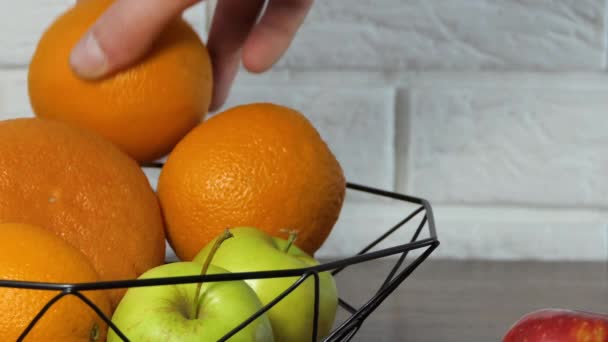 Uma mão coloca frutas em uma cesta de frutas na cozinha. Laranjas de cesto de frutas, maçãs, bananas, toranja — Vídeo de Stock