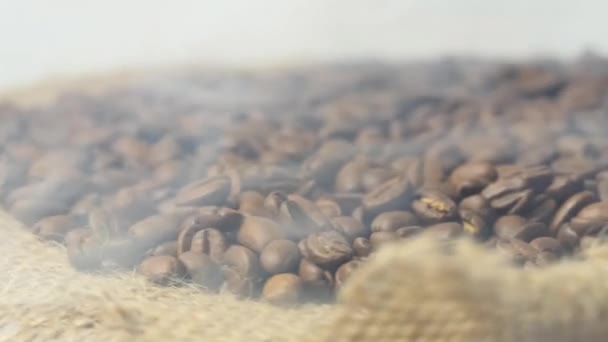 Gros plan sur les grains de café torréfiés. Les grains de café aromatiques sont de la fumée torréfiée provenant des grains de café. Les grains frits reposent sur un sac brun. Au ralenti. Caméra tournante. — Video