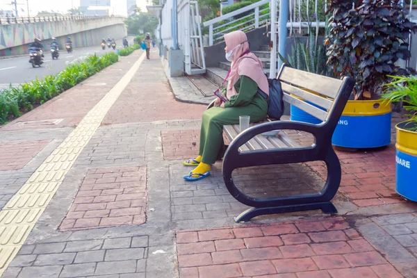 Jakarta, Endonezya, 16 Mart 2022: Asyalı Müslüman bir kadın kaldırımın kenarında uzun bir sandalyede oturuyor, toplu taşıma araçlarının onu işe götürmesini bekliyor..