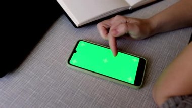The blonde is holding a phone with a green screen. Beautiful hands of a girl leaf through the tape on the phone against the background of a notebook. New technology concept. To advertise a new app.