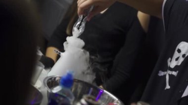 Siyah tişörtlü, kafatası olan tanınmayan bir adamın eli pens kullanır ve dumanlı bir şişeye buz koyar. Kapağı kapalı bir şekilde kapatır. Karanlık laboratuvarda zehir deneyleri yapan insanlar..