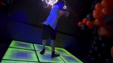 Beyaz tişörtlü, siyah şortlu küçük mutlu çocuk gece kulübünde dans ediyor. Arka planda parlayan neon levha partisi, duvarlarda örümcek ağı. Cadılar Bayramı konsepti.