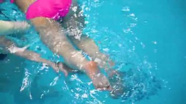 Close up top view of little girl legs hit blue water in swimming pool, splash. Child with mother learns to swimming and floating. Concept of active leisure.