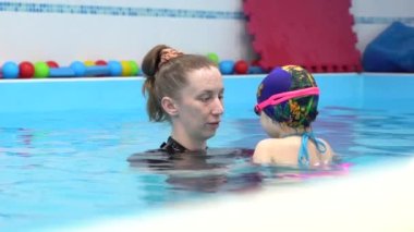 Caucasian woman coach and mom teaches a little child to swim, floating and breathe underwater, gives instructions. Girl wear pink goggles, cap on background of blue water in pool. Indoor view. 
