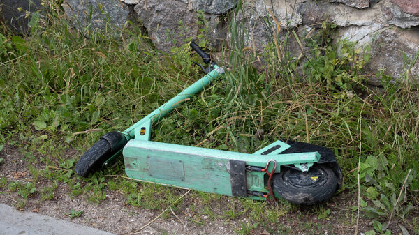 Green electric scooter knocked down in the city on the ground by the fence in the grass. Ecological transportation in the urban environment.