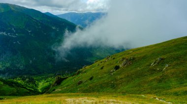 Mountains in clouds on a summer day. Aerial view of mountain top with green trees in fog. Beautiful landscape with high mountains. A mountain ridge separates the cloud from threatening storm clouds.