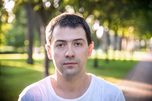 Açık havada, beyaz tişörtlü, doğa parkının arka planında güzel, ciddi bir adamın portresini çek. Moda modeli.