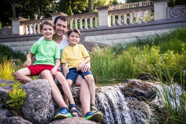 Baba ve iki çocuk, küçük oğlan ve şelale geçmişi olan açık hava portresi. Kristal deresinin yanındaki büyük kayanın üzerinde..