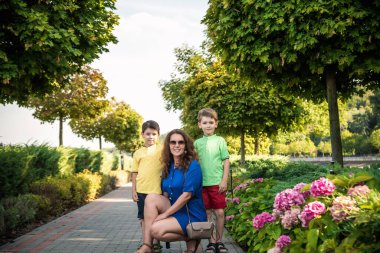 Genç bir kadın çocuklarıyla parkta oynarken eğleniyor. İlkokul çağındaki çocuklar. Aile pitoresk bitkilerle çevrili. Açık hava oyunları herkes için kullanışlı.