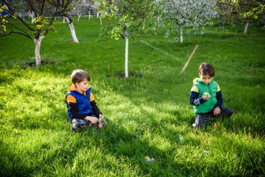 Paskalya yumurtası avındaki çocuklar bahar bahçesinde. Çiçek çayırında renkli yumurta arayan çocuklar. Küçük çocuk ve kardeşi arkadaşı çocuk dışarıda oynuyorlar..