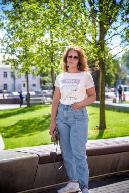 Şehir arka planında günlük kıyafetler, güneş gözlüğü ve kot pantolon giyen esmer bir kızın yakın plan portresi. Sevimli bir bayan şehrin caddesinde dikilip doğrudan kameraya bakıyor..