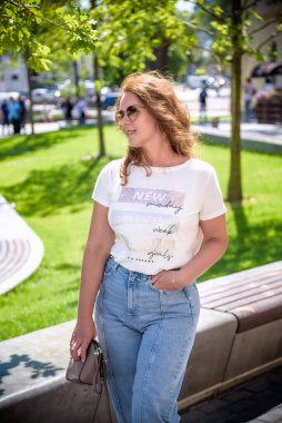 Şehir arka planında günlük kıyafetler, güneş gözlüğü ve kot pantolon giyen esmer bir kızın yakın plan portresi. Sevimli bir bayan şehrin caddesinde dikilip doğrudan kameraya bakıyor..