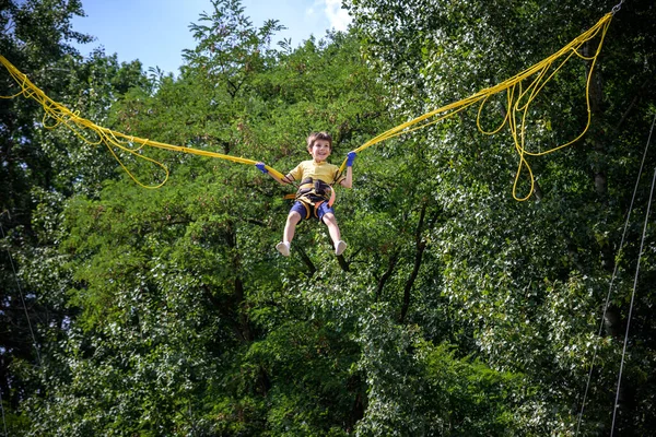 那男孩跳在蹦床上 一个有保险和可伸展的橡皮筋的孩子挂在天空中 快乐童年的概念与游乐园中的游戏 — 图库照片