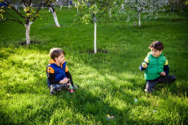 Paskalya yumurtası avındaki çocuklar bahar bahçesinde. Çiçek çayırında renkli yumurta arayan çocuklar. Küçük çocuk ve kardeşi arkadaşı çocuk dışarıda oynuyorlar..