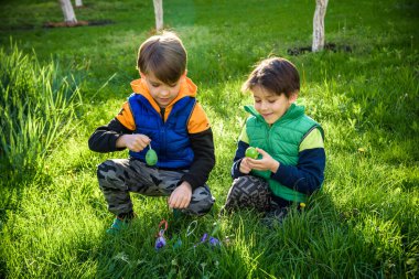 Paskalya yumurtası avındaki çocuklar bahar bahçesinde. Çiçek çayırında renkli yumurta arayan çocuklar. Küçük çocuk ve kardeşi arkadaşı çocuk dışarıda oynuyorlar..