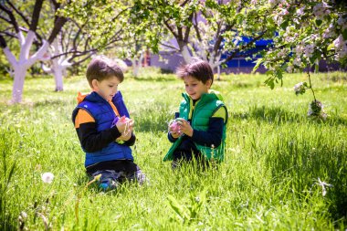 Paskalya yumurtası avındaki çocuklar bahar bahçesinde. Çiçek çayırında renkli yumurta arayan çocuklar. Küçük çocuk ve kardeşi arkadaşı çocuk dışarıda oynuyorlar..