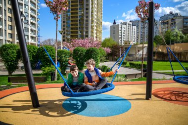 Cheerful boy with his toddler brother swinging in the swing-nest. Entertainment for preschoolers outdoors. Relaxation friendship concept.