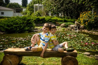 İki erkek kardeş, barışçıl göl kenarındaki parkta bir bankta Lily Nymphaeum 'la oturuyorlar. Çocuklar sıcak yaz gününde doğada dinleniyorlar..