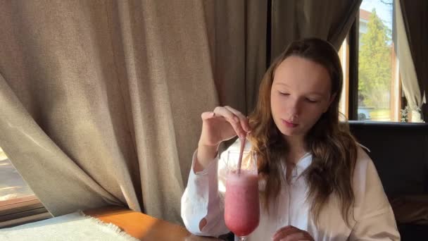 Een meisje drinkt een roze cocktail uit een rood rietje in een restaurant — Stockvideo