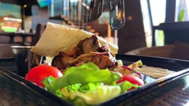 Marul ve domatesli bir restoranda, siyah bir tabakta sıcak kebap ince bir pide ekmeğinin üzerinde duruyor ve çok iştah açıcı görünüyor.