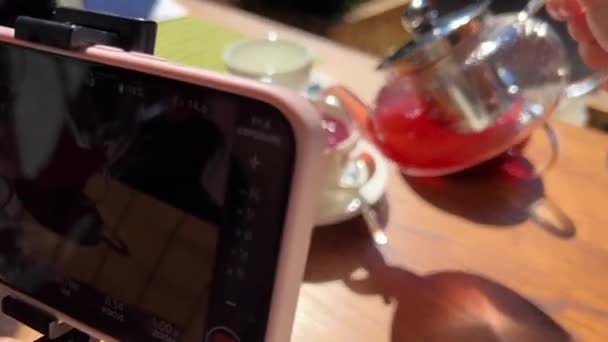 Girl shoots videos on mobile as hot cranberry and currant tea two teapots stand on a wooden table from them poured into a white cup steaming drink — Stock Video