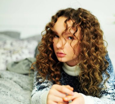 Sıcak, mavi beyaz kazaklı kıvırcık kız gri, sıcak bir battaniyeyle yatağa uzanır. Vazosunun arkasında, yastığın yanında güzel ve dalgın bir fotoğraftır.