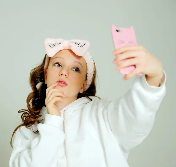 Adolescente em um capuz branco no estúdio em um fundo branco várias caretas, ela é jovem ela tem um telefone celular em suas mãos ela toma uma selfie e admira a si mesma e uma bandagem rosa — Fotografia de Stock