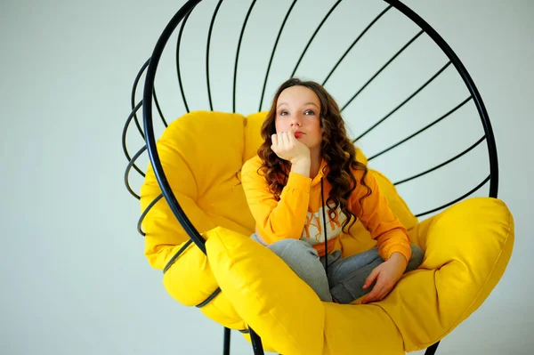 Cadeira de balanço uma menina em uma camisola amarela brilhante senta-se em uma cadeira amarela suculenta e contorce-se enfrenta emoções diferentes no fundo branco do estúdio adolescente está se divertindo e ela está sorrindo — Fotografia de Stock