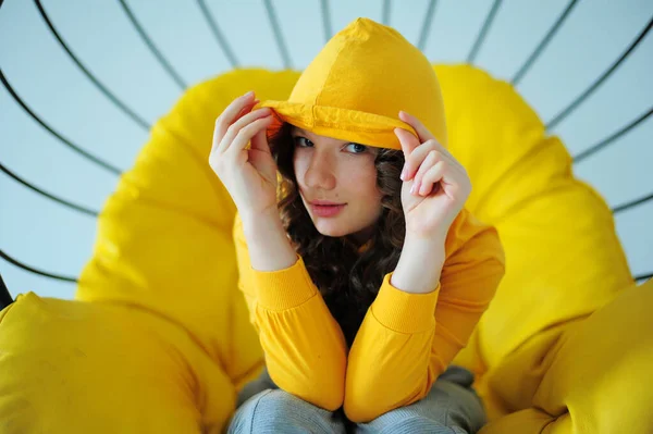 Cadeira de balanço uma menina em uma camisola amarela brilhante senta-se em uma cadeira amarela suculenta e contorce-se enfrenta emoções diferentes no fundo branco do estúdio adolescente está se divertindo e ela está sorrindo — Fotografia de Stock