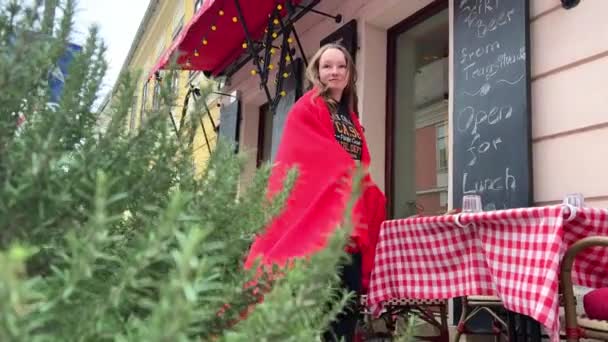 Uma adolescente em uma camisola preta se envolve em um cobertor vermelho brilhante e se senta em uma mesa com uma toalha de mesa em uma gaiola 05.04.22 Pest-Buda café em Budapeste Hungria — Vídeo de Stock