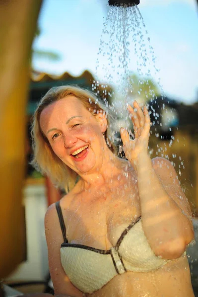 A grown woman laughs in the shower outside squinting one eye cheerfully peeking out from behind the water in a white swimsuit