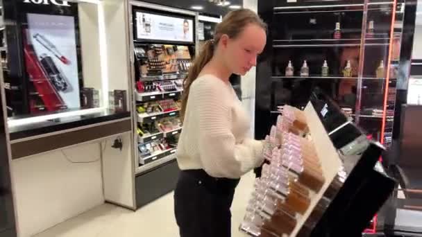 Teenage girl in white jacket and black pants walks around cosmetics store looks at lipstick lip gloss 06.04.22 Paris France — 비디오