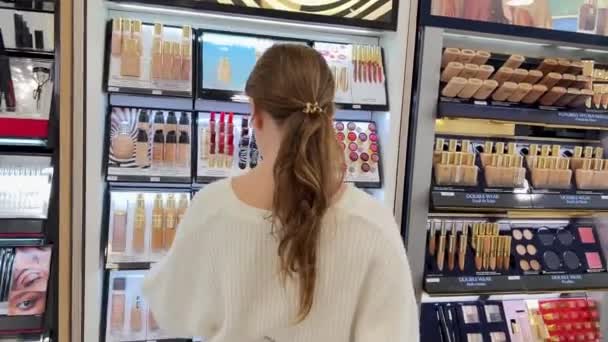 Teenage girl in white jacket and black pants walks around cosmetics store looks at lipstick lip gloss 06.04.22 Paris France — 비디오