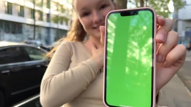 Nastoletnia dziewczyna w beżowej kurtce trzyma telefon z chromatą i bardzo zadowolona wzrusza ramionami i uśmiecha się — Wideo stockowe