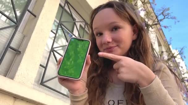 フレーム内の十代の女の子は非常にかわいい笑顔は、携帯電話の画面を示し、画面とクラスに揺れるページをオンにします — ストック動画