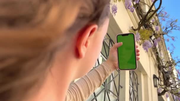 Girl looks at the phone lifting it up with chromate blonde natural hair developing green screen flipping her finger against the background of a beige building — Vídeo de Stock