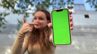 Gevşek saçlı, kahverengi saçlı genç kız, yeşil perdenin arka planında telefonla dans ediyor.