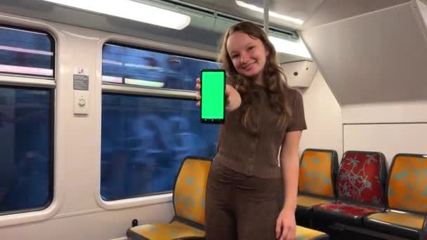 A teenager girl holds a green screen with a chroma key in her hands, dances, smiles, looks into the frame, shows class and is very pleased she is alone in the train rides sunny on the street — Vídeos de Stock