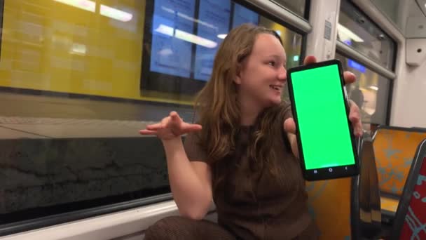 Ein Teenager-Mädchen hält einen grünen Bildschirm mit einem Chroma-Schlüssel in der Hand, tanzt, lächelt, schaut in den Rahmen, zeigt Unterricht und ist sehr froh, dass sie allein in der Bahn ist. — Stockvideo