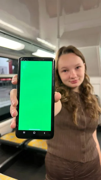 Nastolatka trzyma zielony ekran z kluczem chroma w rękach, tańczy, uśmiecha się, patrzy w kadr, pokazuje klasę i jest bardzo zadowolona, że jest sama w pociągu jeździ słonecznie po ulicy — Zdjęcie stockowe