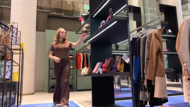 Hermosa adolescente chica camina en una tienda de ropa mirando bolsos sombreros de ropa y varias otras cosas — Vídeo de stock