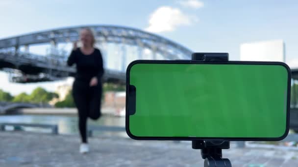 A mobile phone with a green screen in the foreground behind a contented adult woman mom is high on the background of the bridge she jumps dances jumps — Vídeo de stock