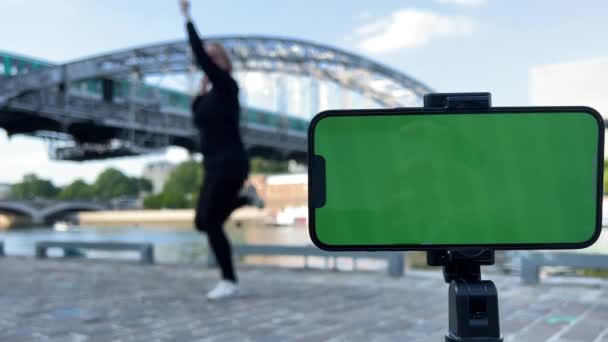 Ein Mobiltelefon mit grünem Bildschirm im Vordergrund hinter einer zufriedenen erwachsenen Mutter ist hoch oben auf dem Hintergrund der Brücke, auf der sie springt, tanzt Sprünge — Stockvideo